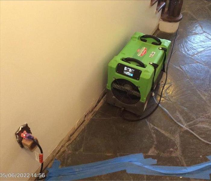 A SERVPRO dehumidifier operating near a wall with removed baseboards and an outlet without a cover