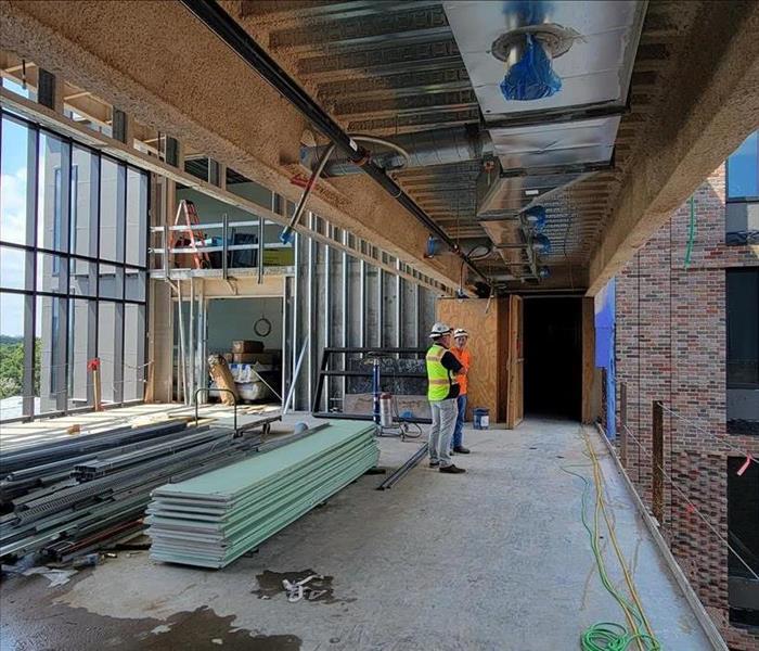 The upper floor of an unfinished construction area with saturated steel and subflooring is visible. Two technicians in safety